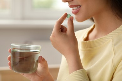 Photo of Young woman with glass of water taking dietary supplement pill indoors, closeup