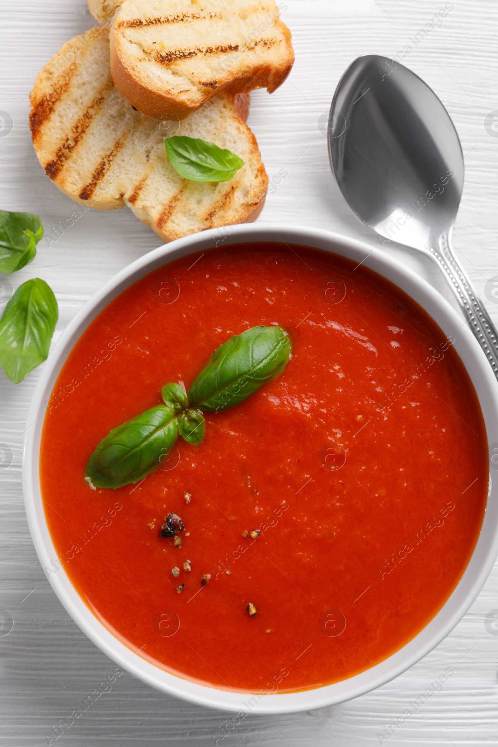 Photo of Delicious tomato cream soup in bowl served with pieces of grilled bread on white wooden table, flat lay