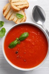 Delicious tomato cream soup in bowl served with pieces of grilled bread on white wooden table, flat lay