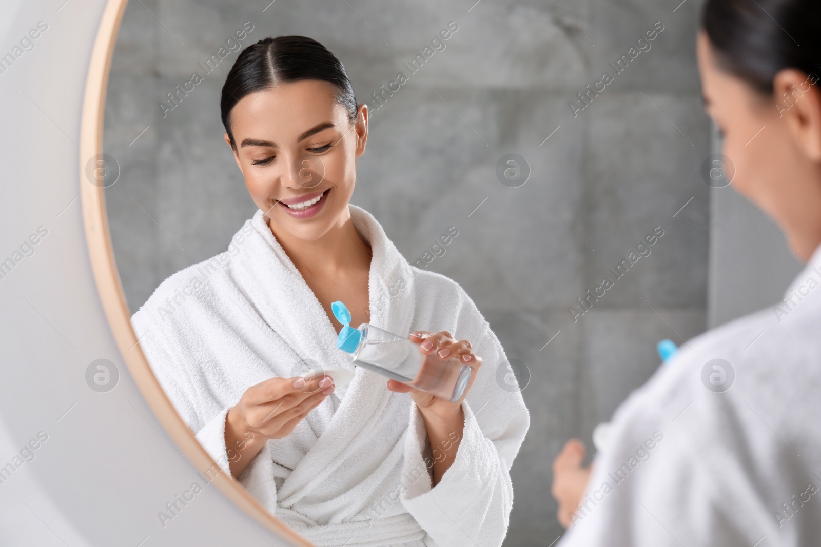 Photo of Beautiful woman pouring makeup remover from bottle onto cotton pad near mirror indoors