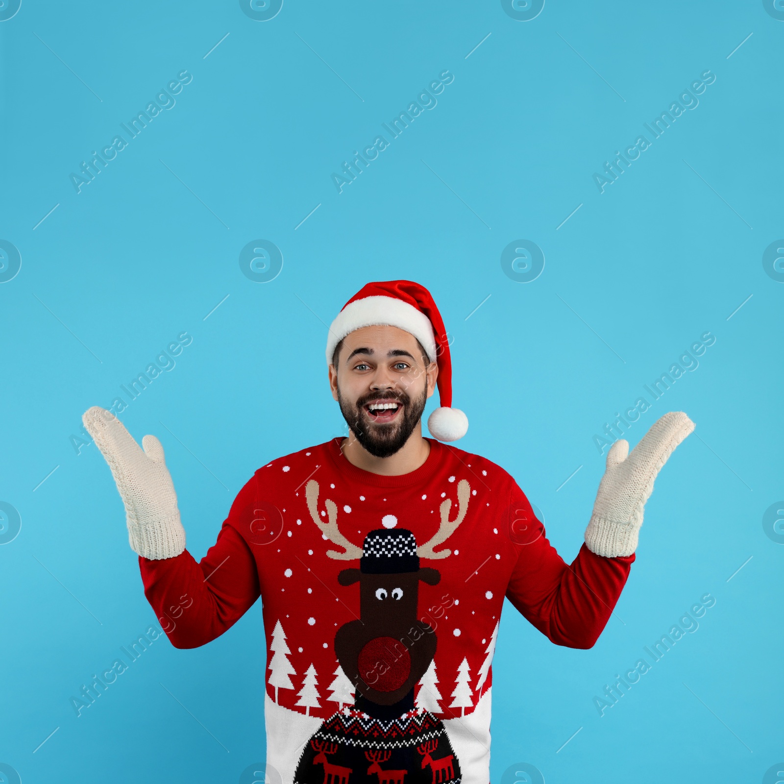 Photo of Happy young man in Christmas sweater, Santa hat and knitted mittens on light blue background