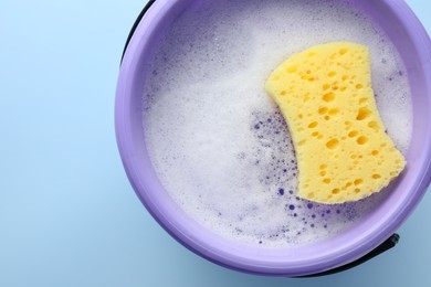 Bucket with foam and sponge on light blue background, top view