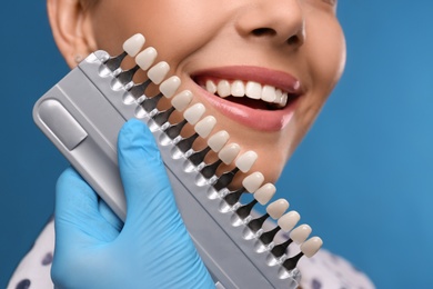Photo of Doctor checking young woman's teeth color on blue background, closeup. Cosmetic dentistry