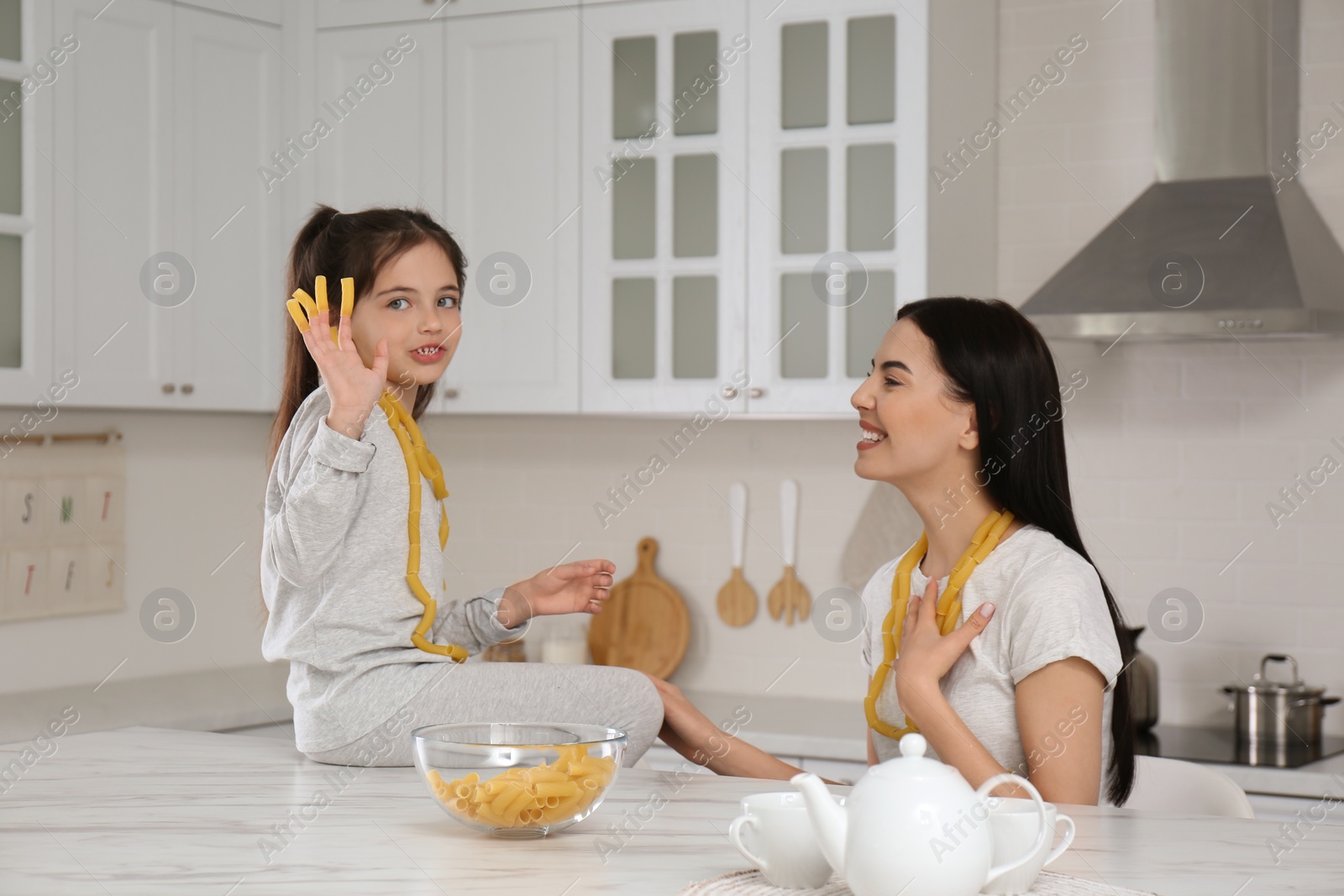 Photo of Young mother and her daughter with necklaces made of pasta having fun in kitchen