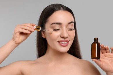 Happy young woman with bottle applying essential oil onto face on light grey background