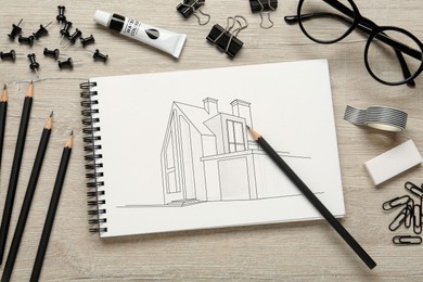Image of Sketch of building in notebook, glasses, pencils and other stationery on wooden table, flat lay