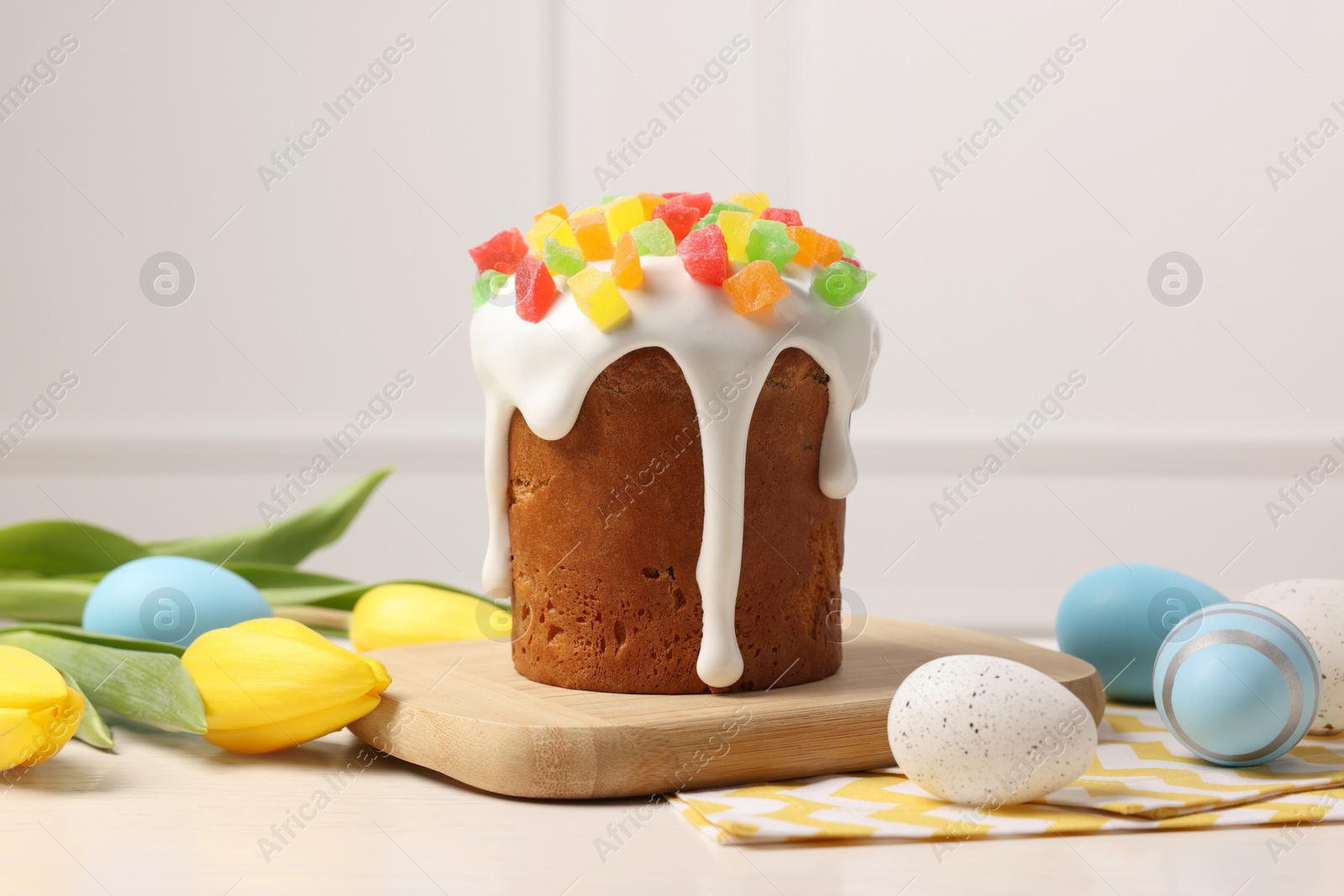 Photo of Delicious Easter cake with pieces of candied fruits, painted eggs and tulips on white table indoors