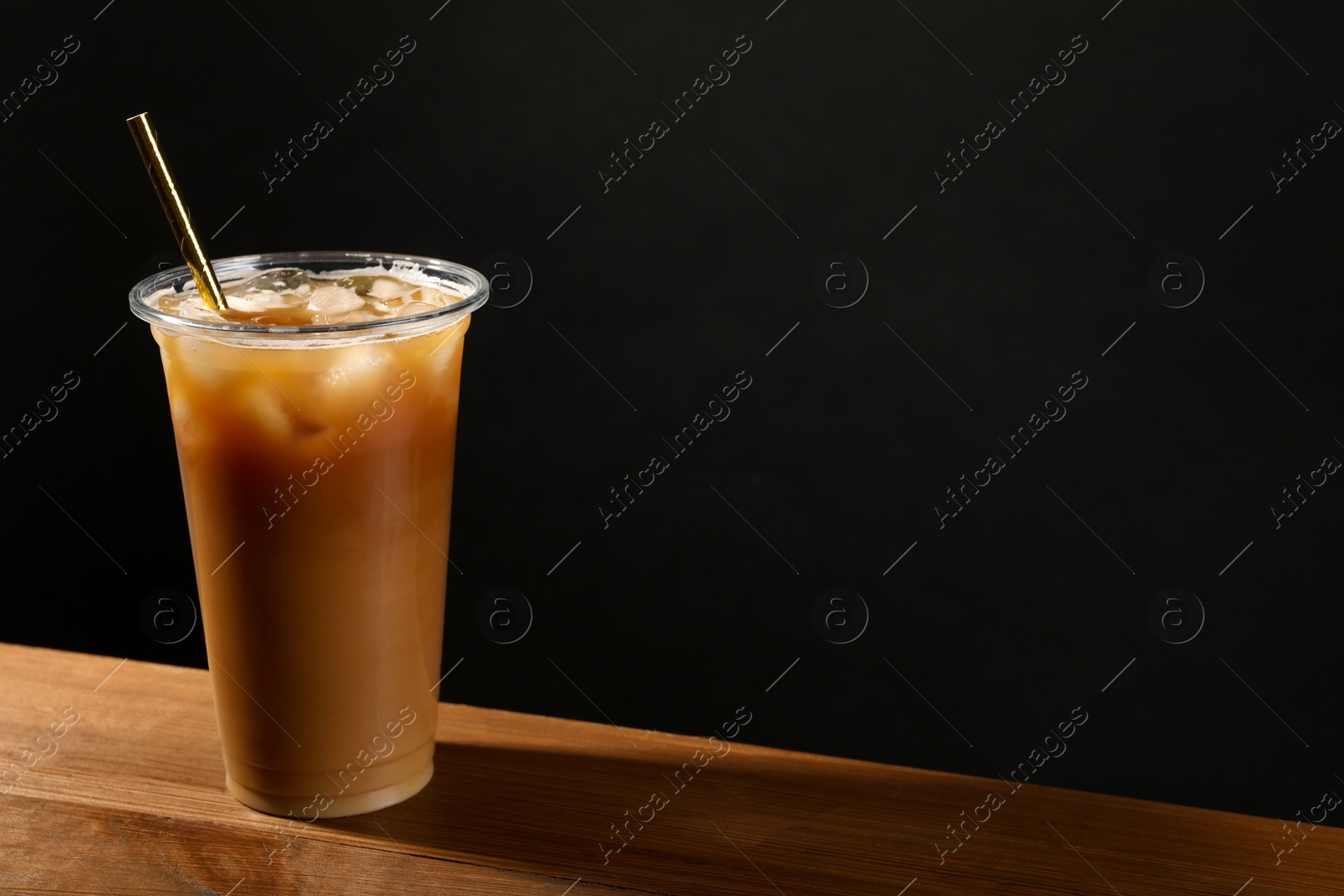 Photo of Refreshing iced coffee with milk in takeaway cup on wooden table against black background, space for text