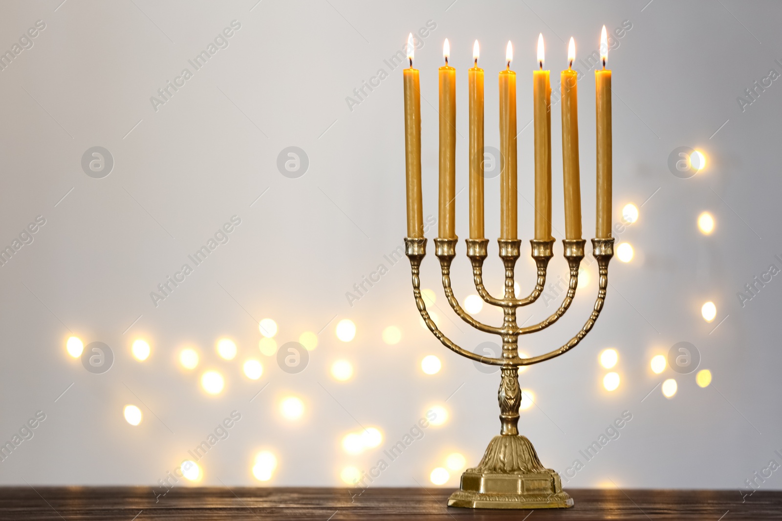 Photo of Golden menorah with burning candles on table against light grey background and blurred festive lights, space for text