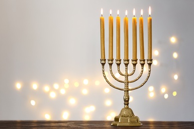Photo of Golden menorah with burning candles on table against light grey background and blurred festive lights, space for text
