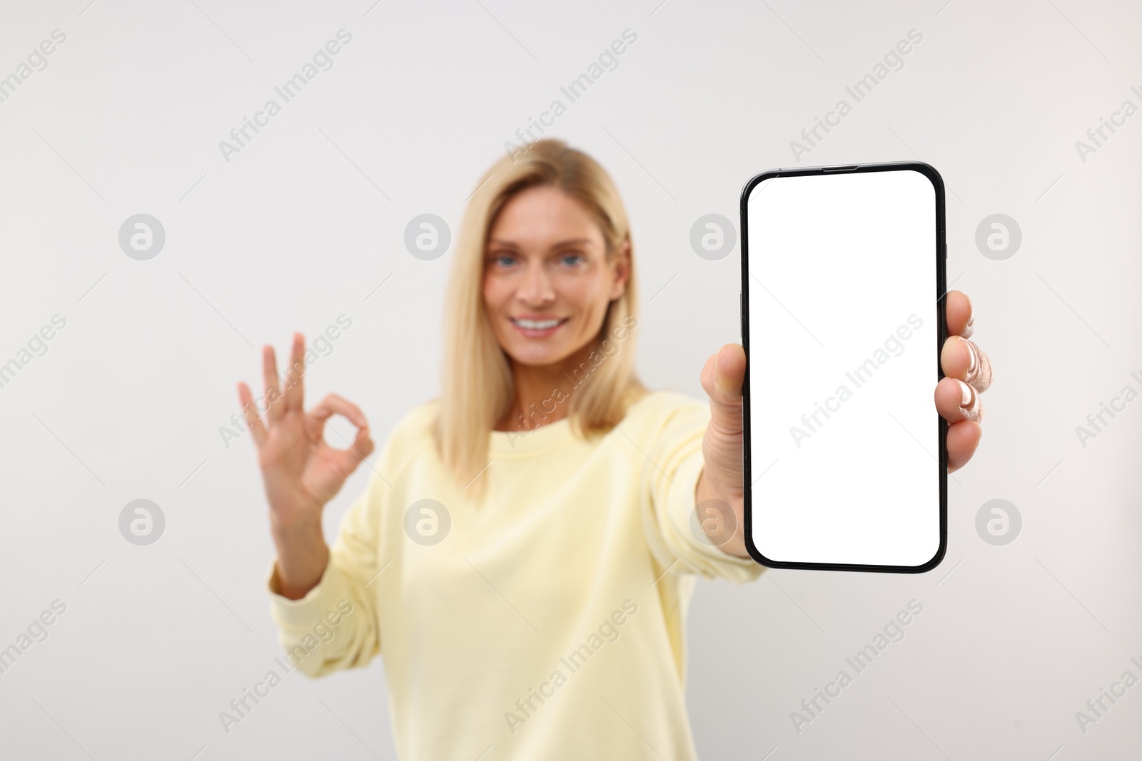 Photo of Happy woman holding smartphone with blank screen and showing OK gesture on white background, selective focus