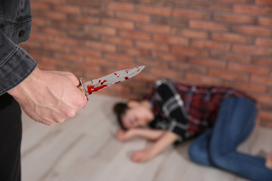 Photo of Man with bloody knife and his victim on floor indoors, closeup. Dangerous criminal