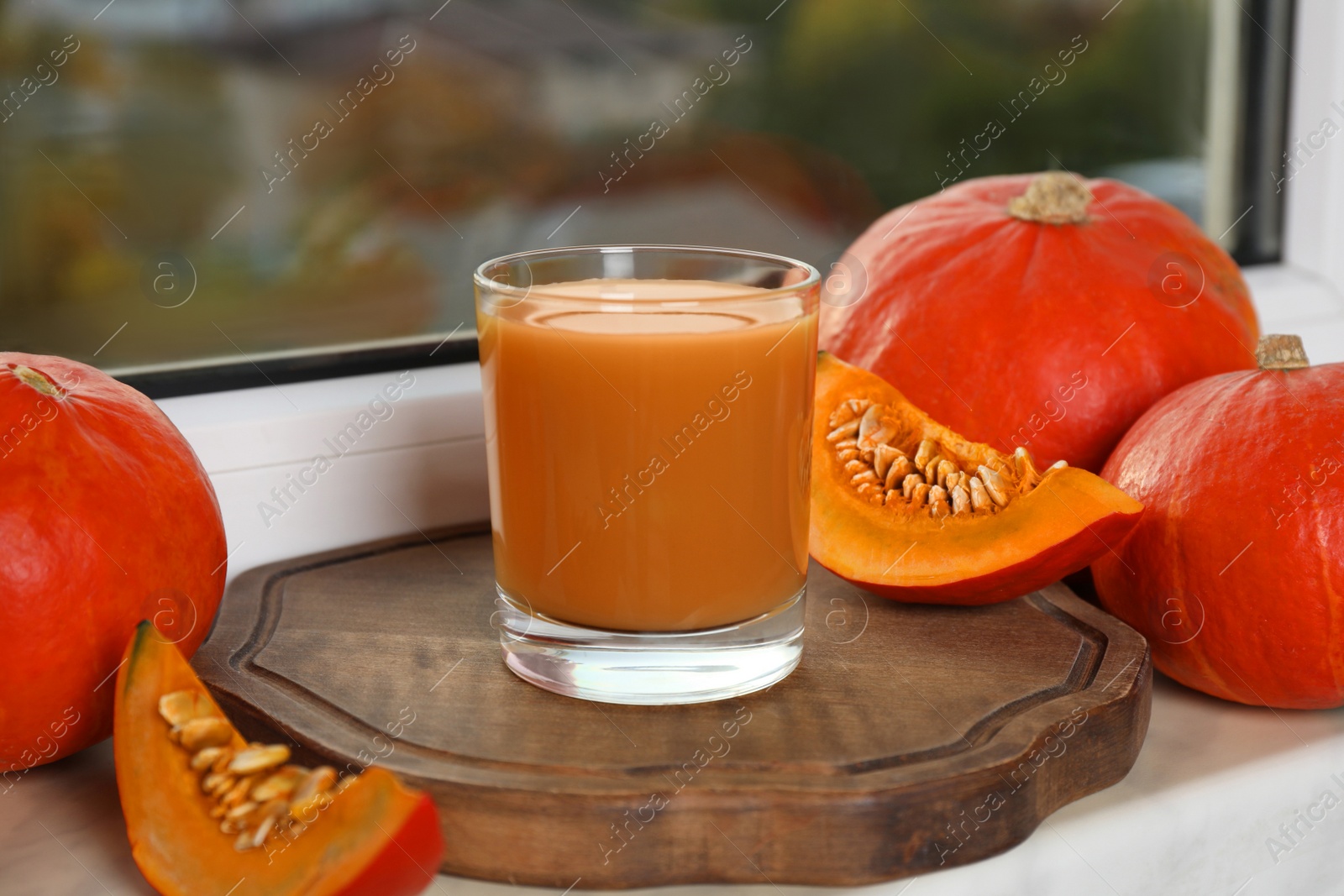 Photo of Tasty pumpkin juice in glass, whole and cut pumpkins on windowsill indoors