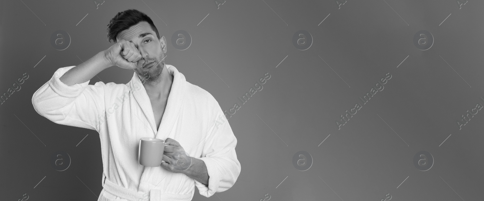 Image of Young man in bathrobe with cup of coffee on grey background, space for text. Black and white photography