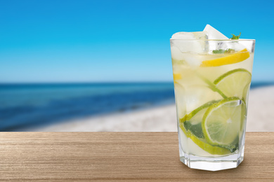 Image of Citrus lemonade with ice cubes on wooden table at beach, space for text