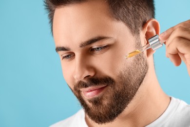 Photo of Handsome man applying cosmetic serum onto face on light blue background, closeup