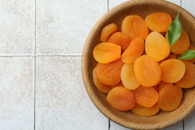 Photo of Wooden bowl of tasty apricots and space for text on white tiled table, top view. Dried fruits