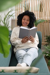 Relaxing atmosphere. Happy woman reading book on sofa surrounded by beautiful houseplants in room