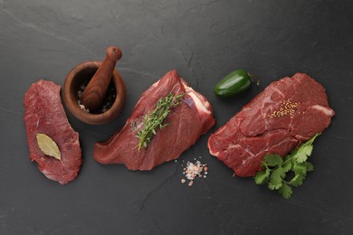 Photo of Pieces of raw beef meat, mortar with pestle and spices on black table, flat lay