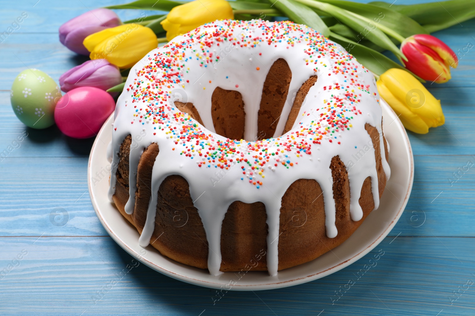 Photo of Glazed Easter cake with sprinkles, painted eggs and tulips on blue wooden table, closeup