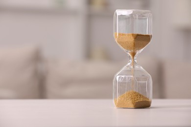 Photo of Hourglass with flowing sand on white table against blurred background. Space for text