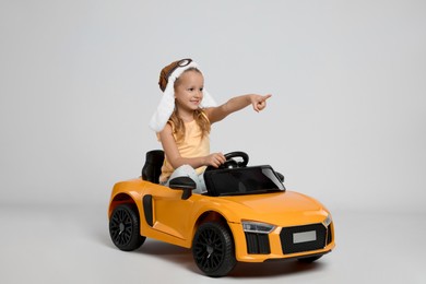 Photo of Cute little girl in pilot hat driving children's electric toy car on grey background