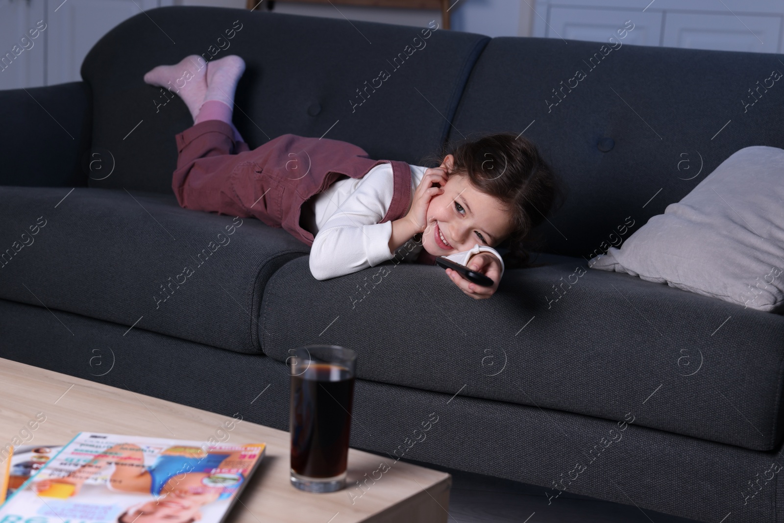 Photo of Cute little girl changing TV channels with remote control on sofa at home