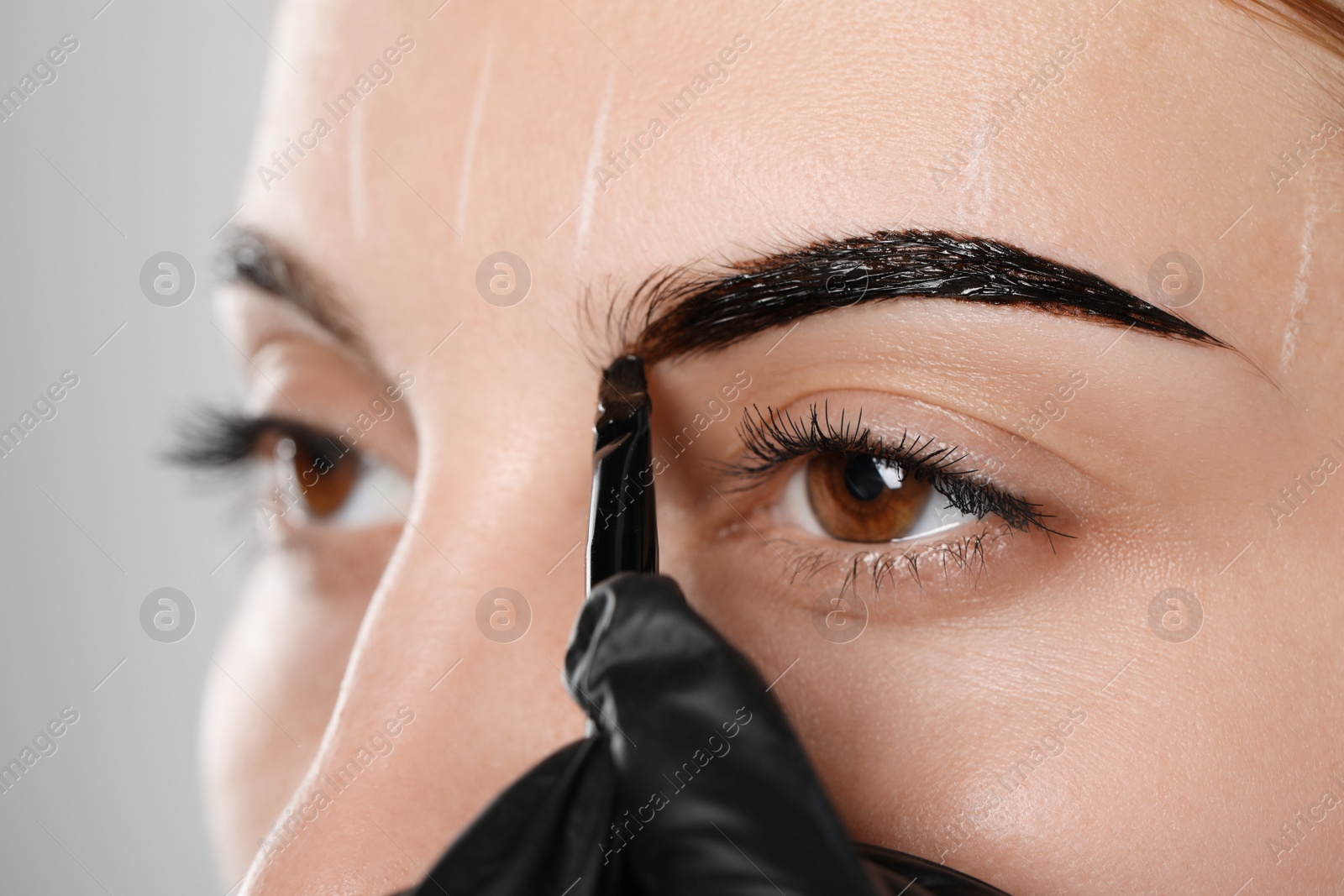 Photo of Beautician applying tint during eyebrows correction procedure on grey background, closeup