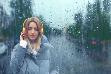 Image of Beautiful woman with headphones listening to music at city street on rainy day, view through wet window