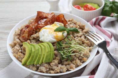 Photo of Delicious boiled oatmeal with poached egg, bacon, avocado and microgreens on table, closeup