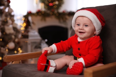 Photo of Cute little baby wearing Santa Claus suit sitting in armchair at home. Christmas celebration