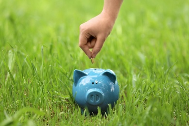 Young woman putting coin into piggy bank on green grass outdoors, closeup