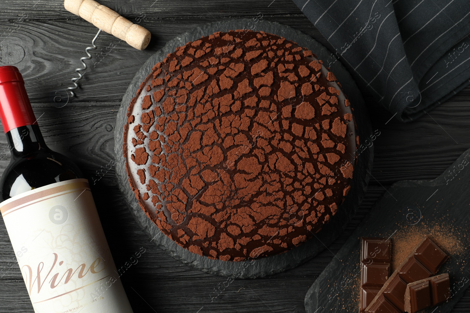 Photo of Delicious truffle cake, bottle of wine, chocolate pieces and corkscrew on black wooden table, flat lay