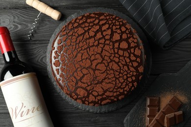 Photo of Delicious truffle cake, bottle of wine, chocolate pieces and corkscrew on black wooden table, flat lay