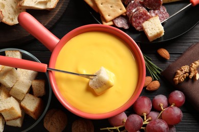 Photo of Pot of tasty cheese fondue and snacks on black wooden table, flat lay