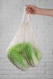 Photo of Woman holding string bag with fresh Chinese cabbages near white brick wall, closeup