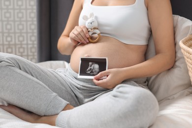 Pregnant woman with ultrasound picture of baby and bunny toy sitting on bed indoors, closeup