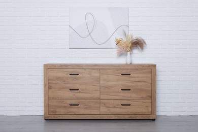 Photo of Wooden chest of drawers, picture and vase near white brick wall. Interior design