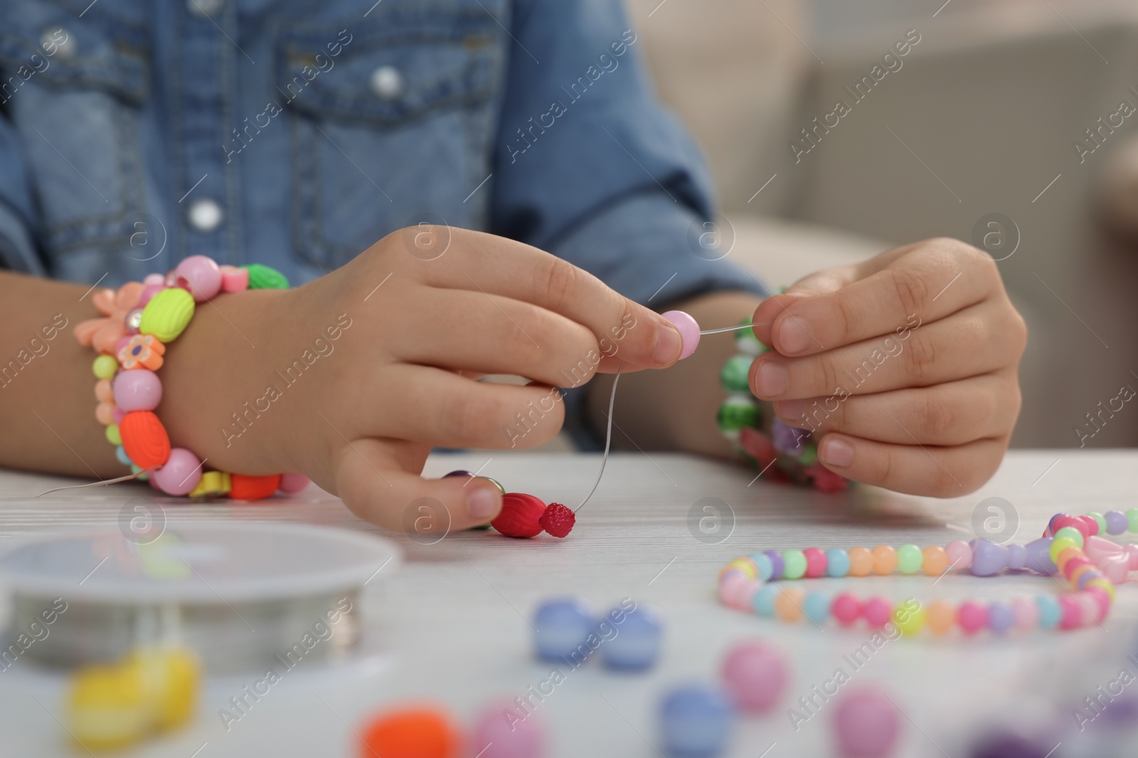 Photo of Little girl making beaded jewelry at table indoors, closeup