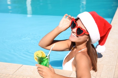 Young woman wearing Santa Claus hat with refreshing drink in swimming pool. Christmas vacation