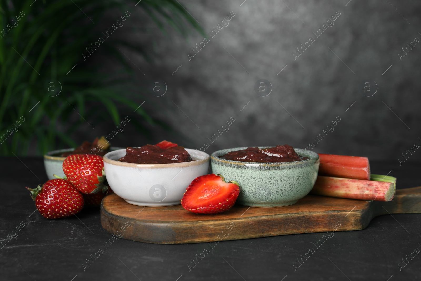 Photo of Tasty rhubarb jam in bowls, stems and strawberries on dark textured table. Space for text