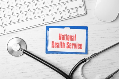 Image of National health service (NHS). Badge with text, stethoscope and keyboard on white wooden background, flat lay