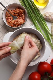 Woman putting uncooked stuffed cabbage roll into ceramic pot at white table, top view