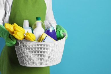 Photo of Spring cleaning. Woman holding basket with detergents, flowers and rags on light blue background, closeup. Space for text