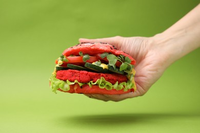 Photo of Woman holding tasty pink vegan burger with vegetables, patty and microgreens on light green background, closeup