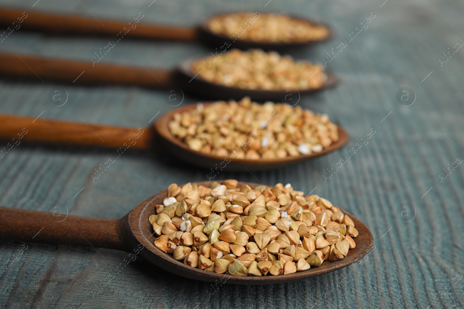 Photo of Uncooked green buckwheat grains in spoons on light blue wooden table