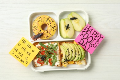 Photo of Lunchbox with fake spider, bugs and Happy Fools' Day note on white wooden table, top view