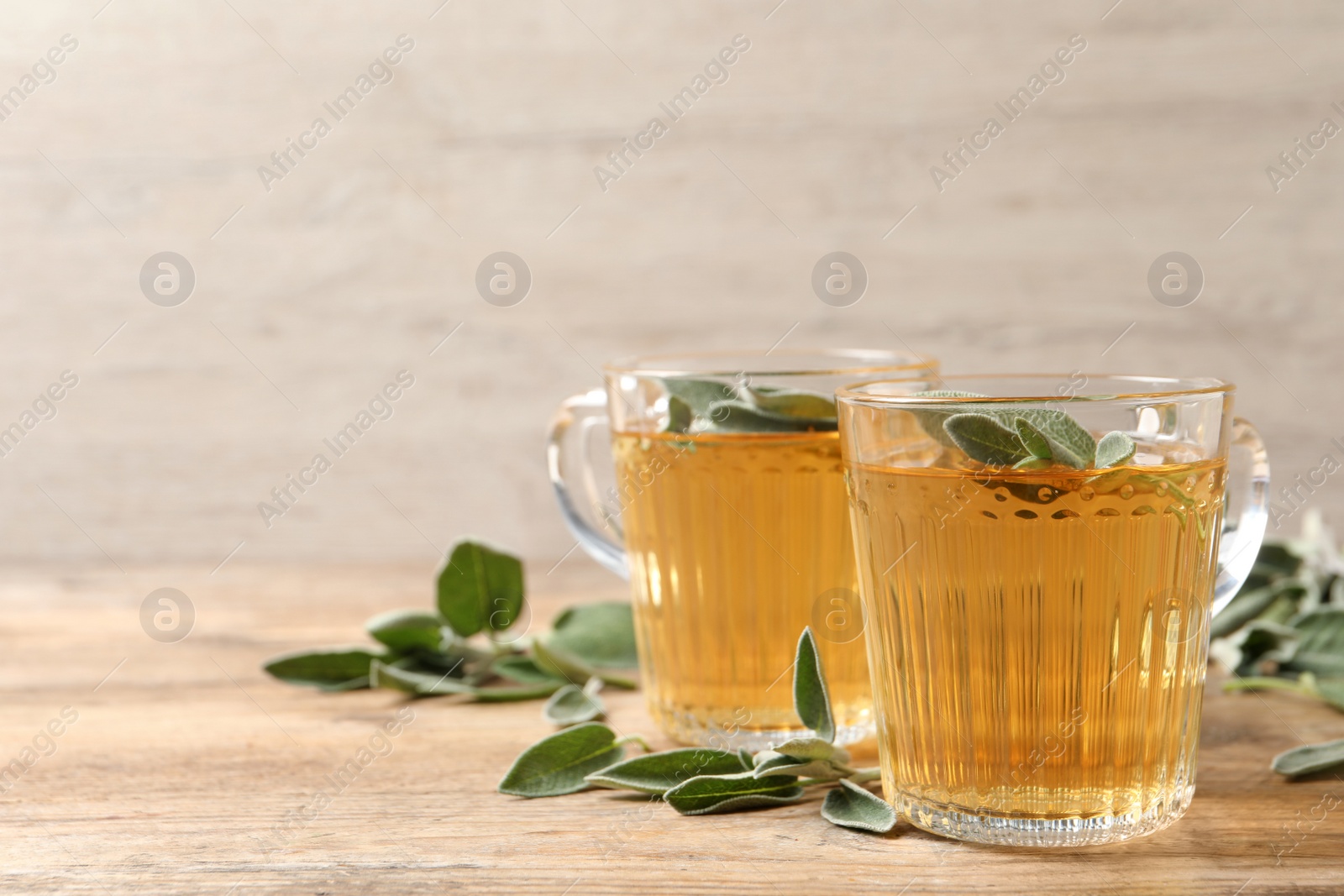 Photo of Cups of aromatic sage tea and fresh leaves on wooden table. Space for text