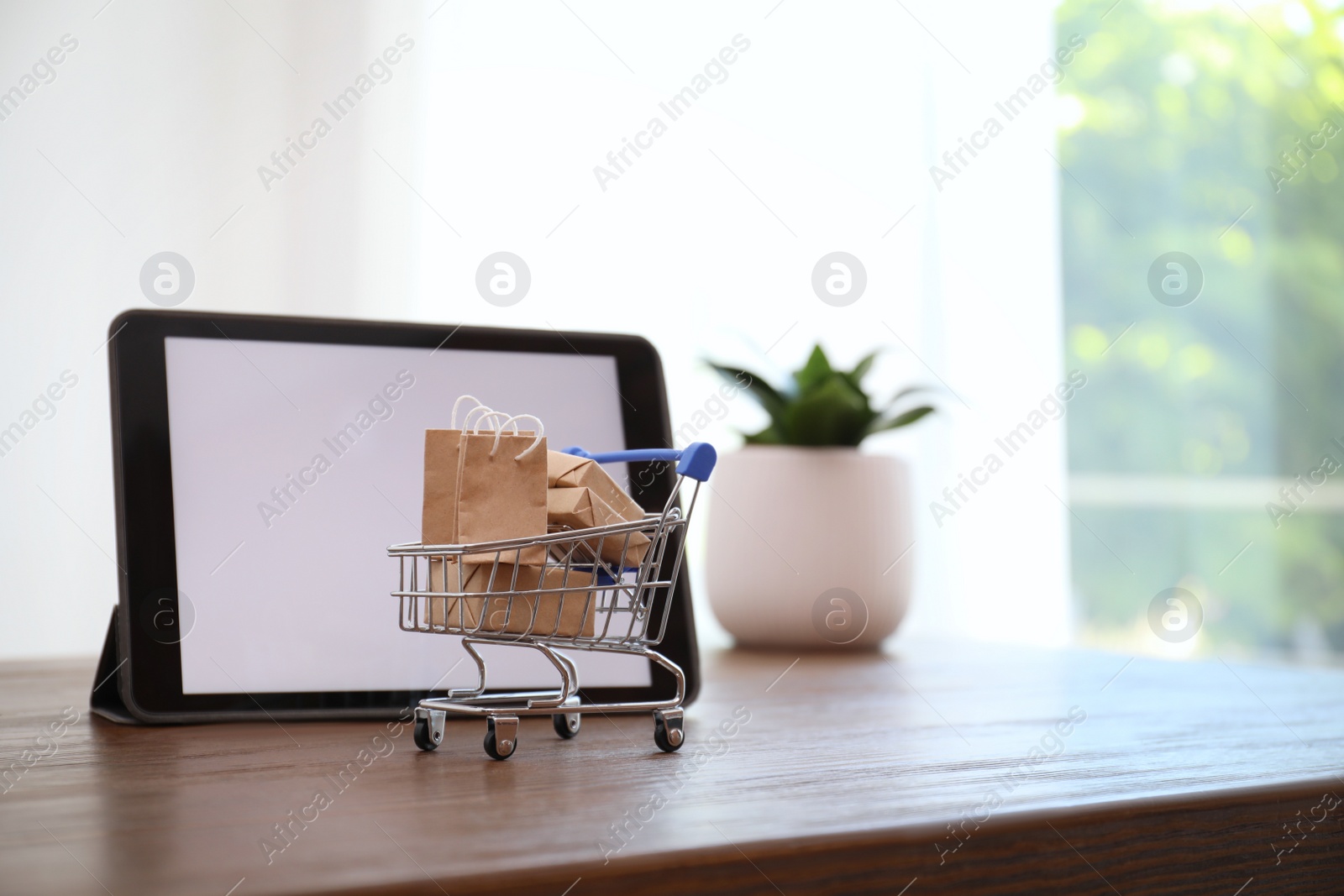 Photo of Internet shopping. Small cart with bags and boxes near modern tablet on table indoors, space for text