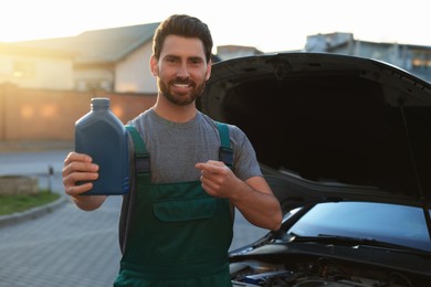Photo of Smiling worker pointing at blue container of motor oil near car outdoors